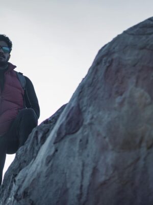 A man with beard and sunglasses hikes on a rocky terrain wearing a red jacket.