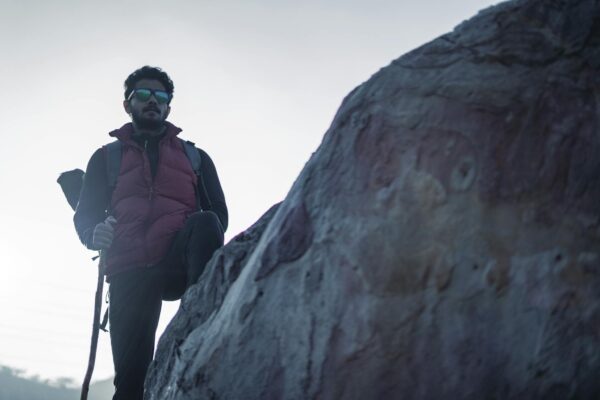 A man with beard and sunglasses hikes on a rocky terrain wearing a red jacket.